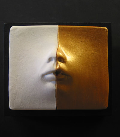Small ceramic wall face mounted on 3.5" square piece of wood painted black. There is a line down the center of the face and one side is natural white and the other painted gold.